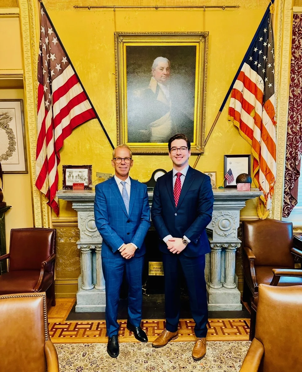 Robert Croak and Austin Hankwitz standing together in suits between two American flags