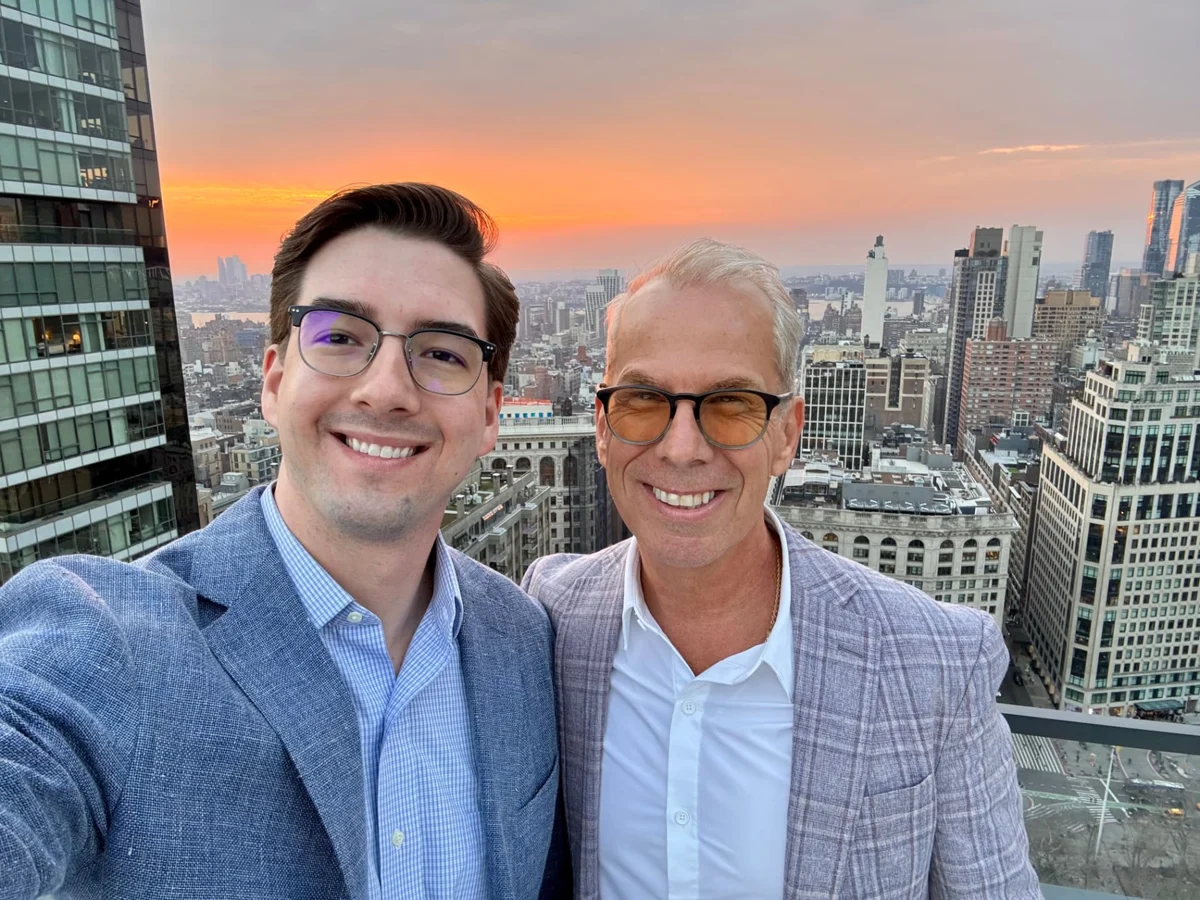 Austin Hankwitz and Robert Croak smiling on a rooftop above the New York City skyline at sunset